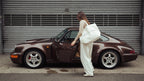 Frau in weißem Outfit trägt eine Studio Élancé Tote Bag neben einem klassischen Porsche 911 vor einem Garagentor